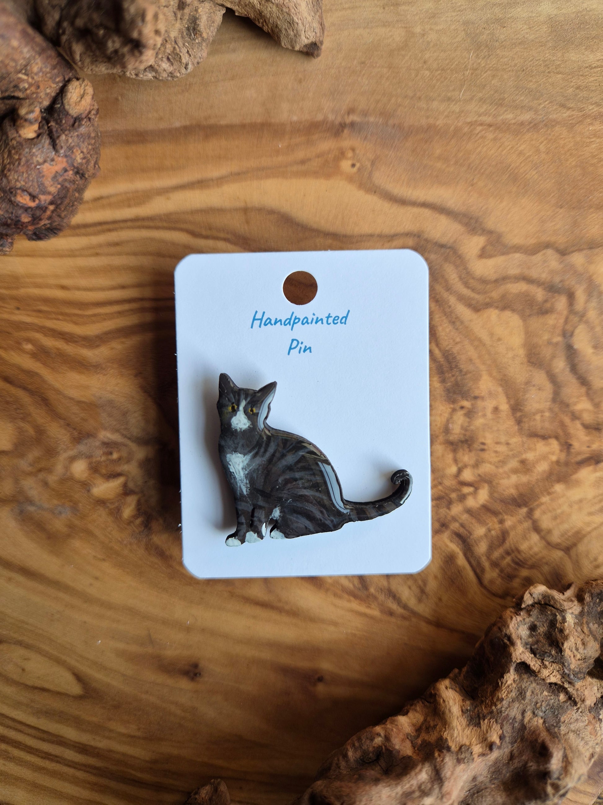 Hand-painted wooden pin badge of a dark tabby coloured cat with white face, chest and paws