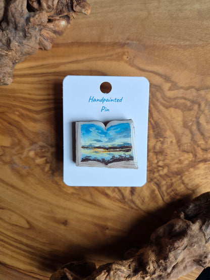 Hand-painted pin badge of an open book shape with a view of a sunset over Findhorn Bay painted inside, painted onto wood