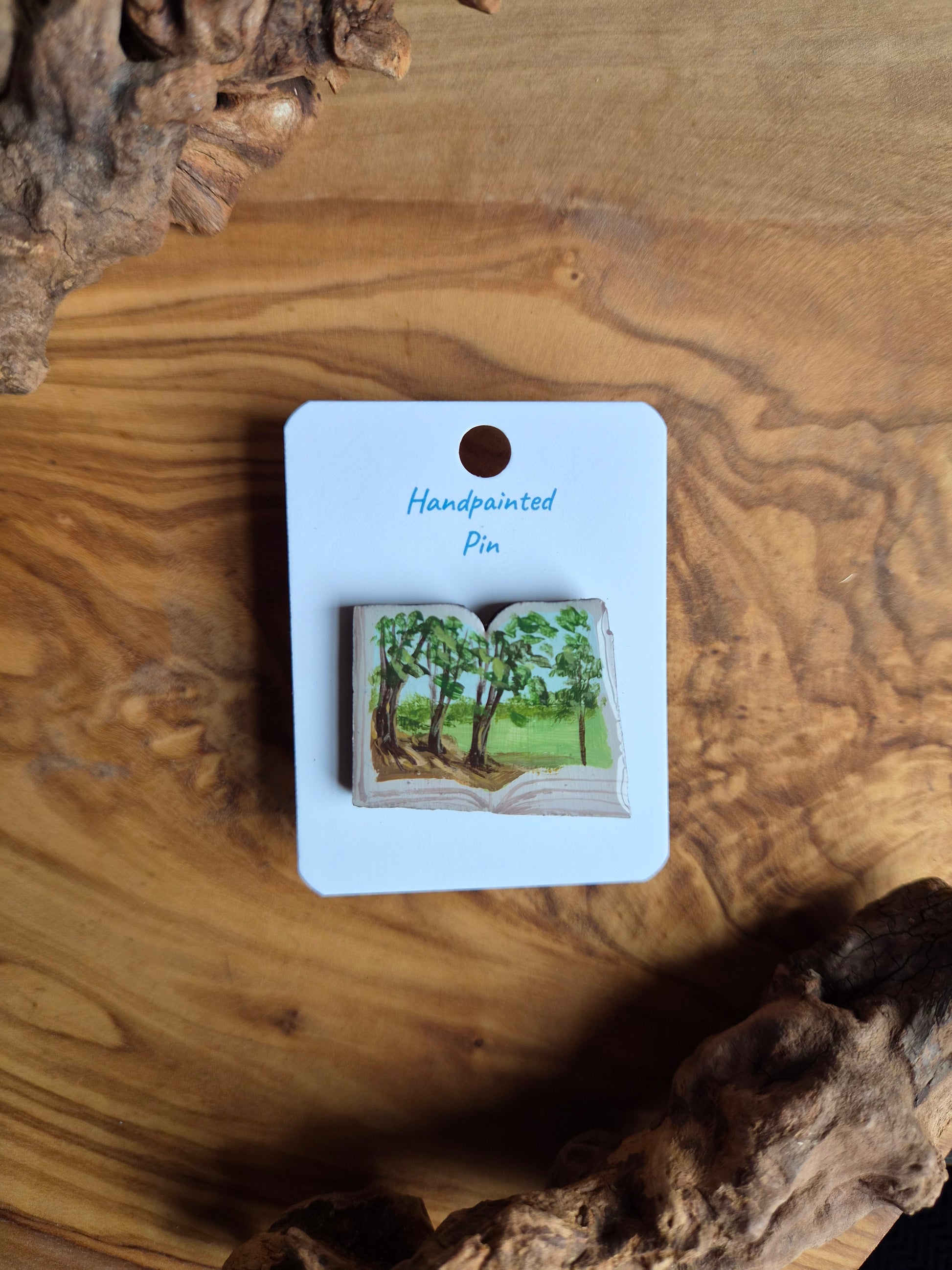 Hand-painted pin badge of an open book shape with a view of a forest with golden hour light filtering through the leaves onto the ground painted inside, painted onto wood