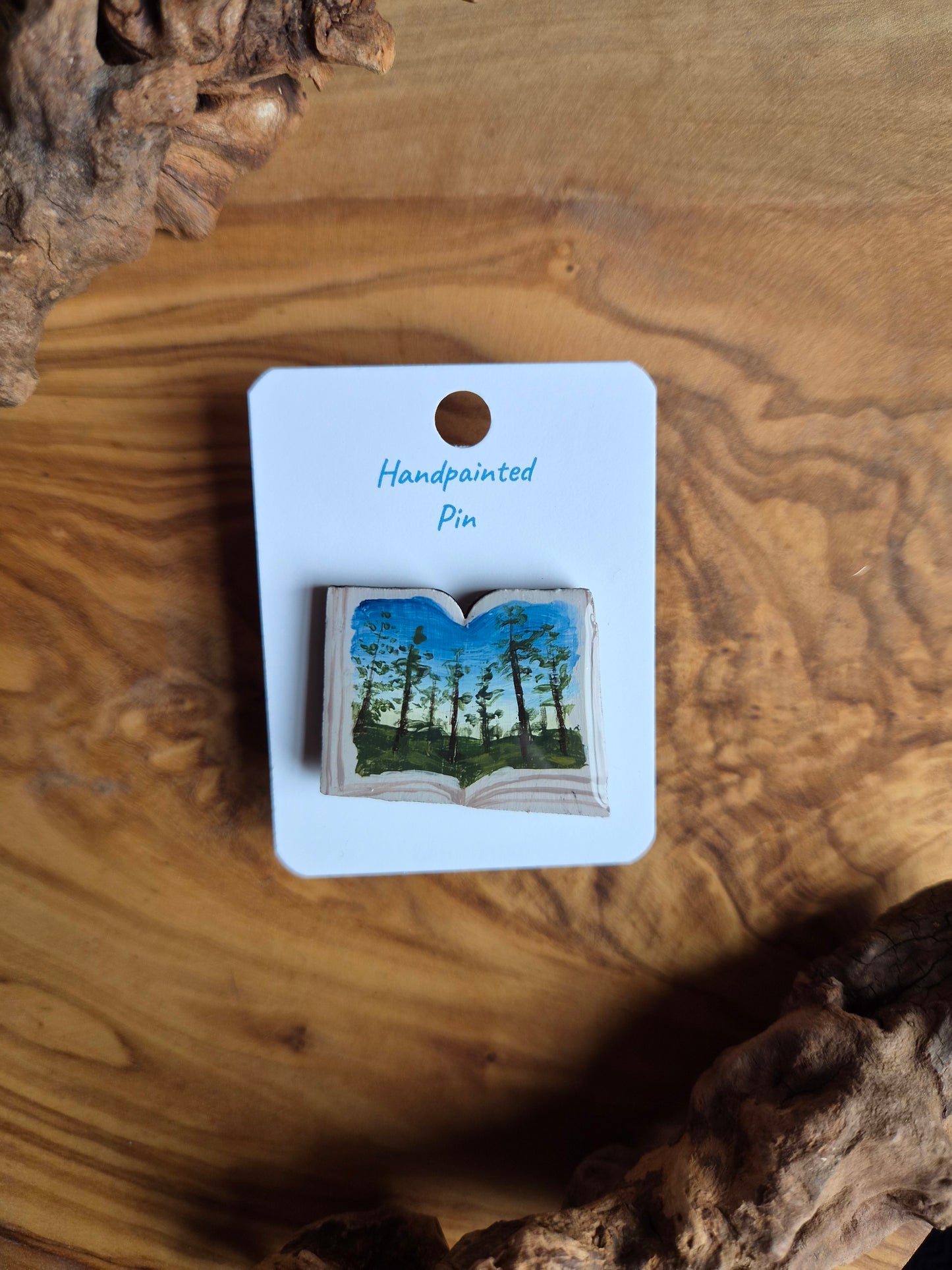Hand-painted pin badge of an open book shape with a view of a forest at dusk in the evening painted inside, painted onto wood