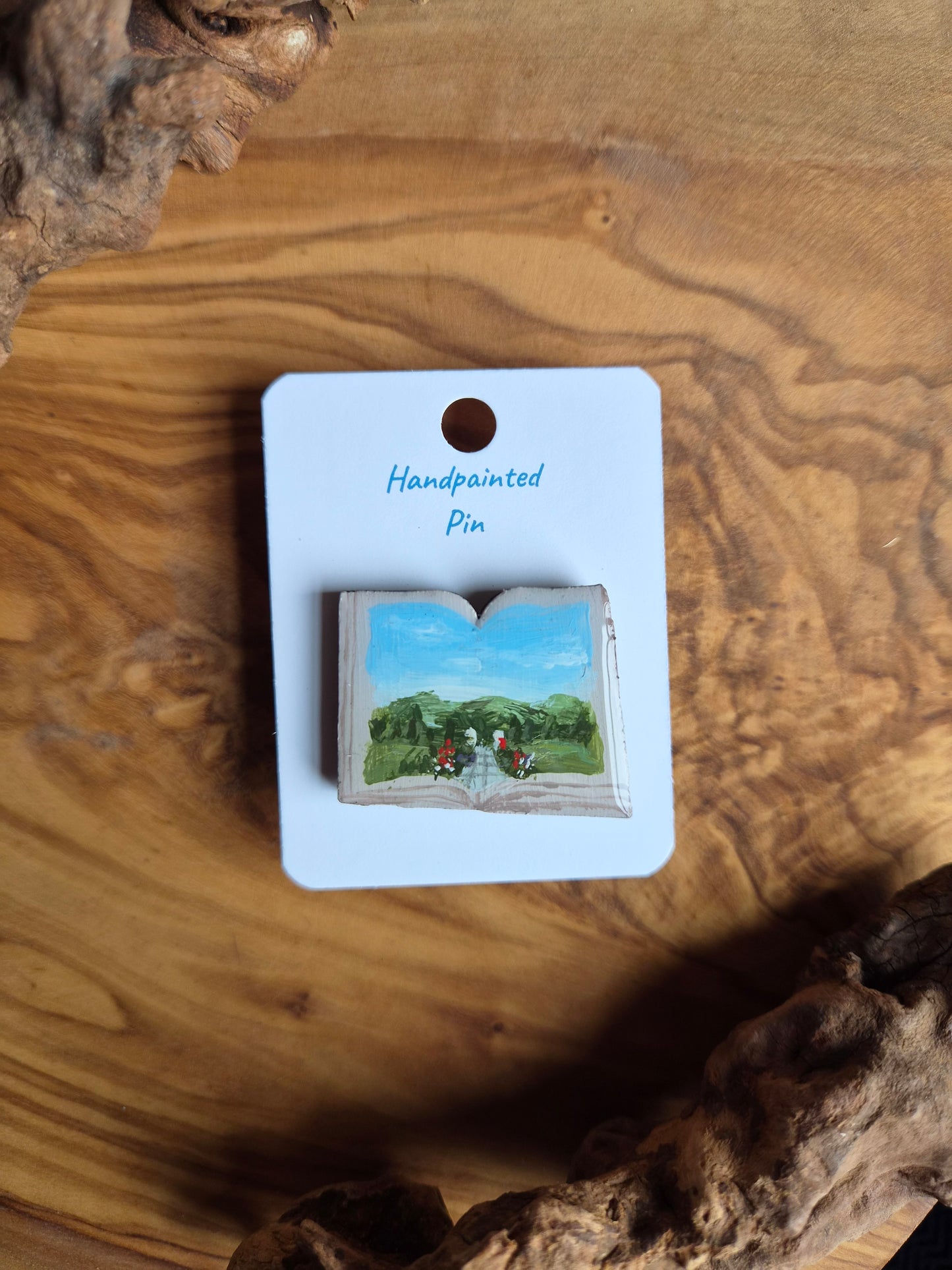 Hand-painted pin badge of an open book shape with a view of a path lined with flowers at Achnagairn Castle painted inside, painted onto wood