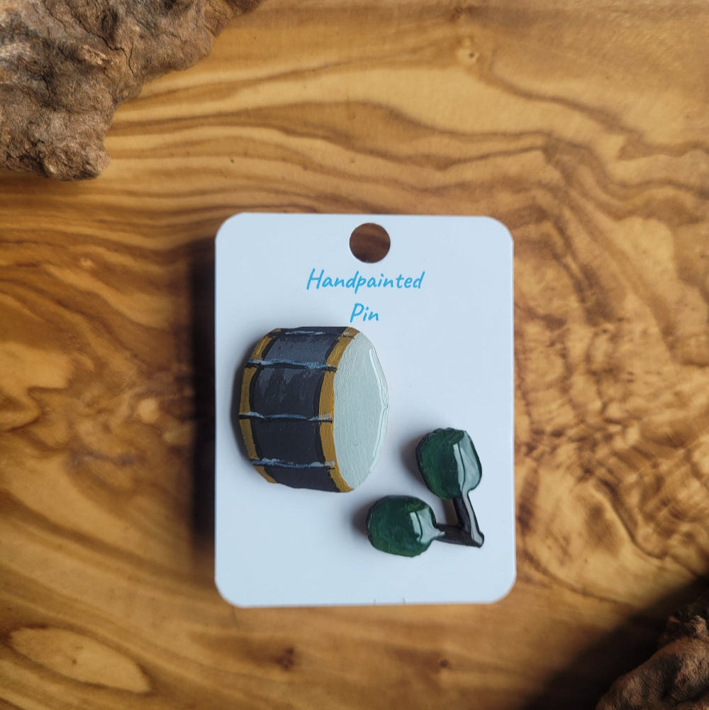 Painted wooden pin of a bass drum and sticks on a card with 'Handpainted pin' text, displayed on a wooden surface.